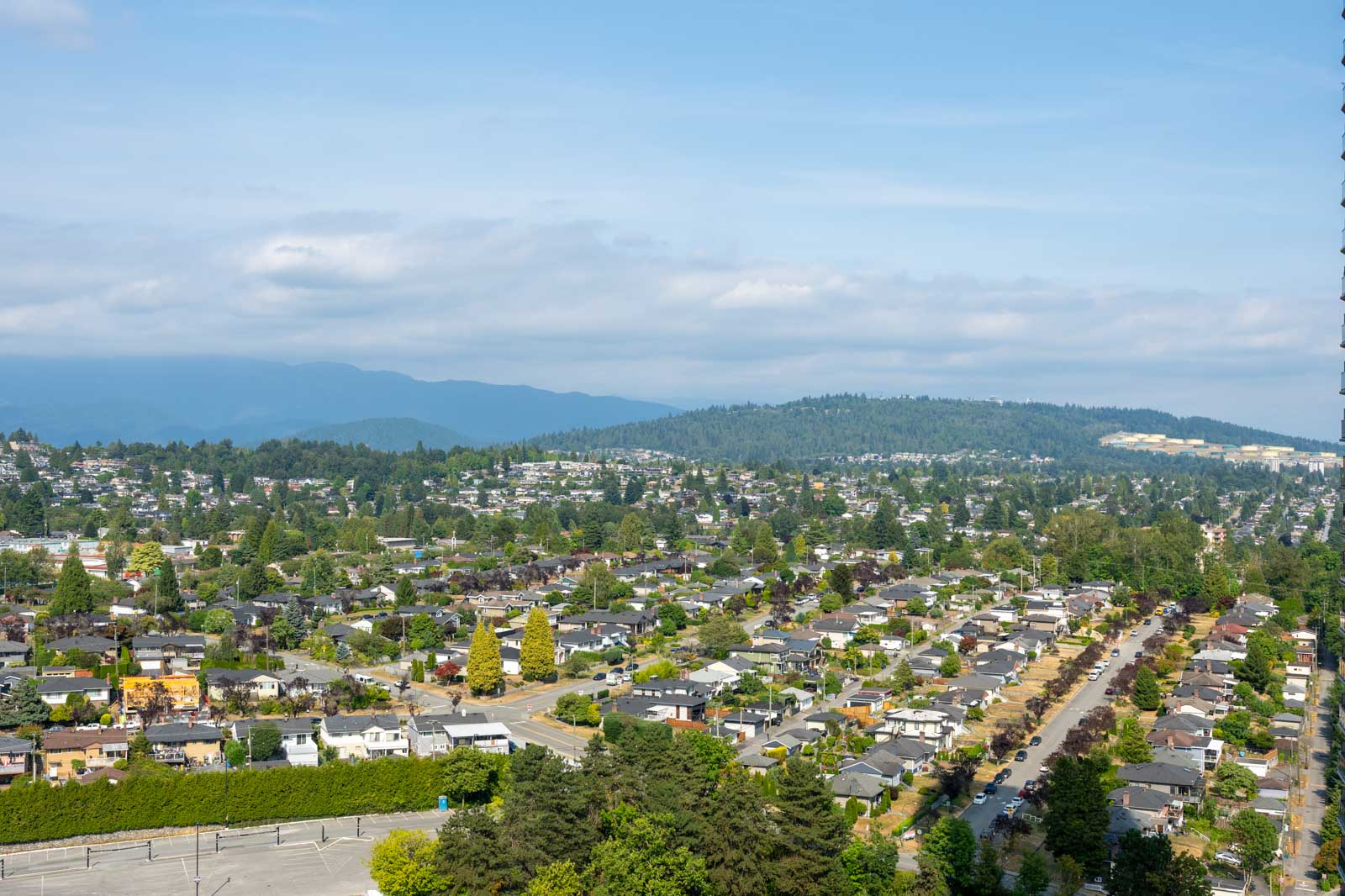 Aerial view of a suburban neighborhood with rows of houses, tree-lined streets, and forested hills in the background under a partly cloudy sky.