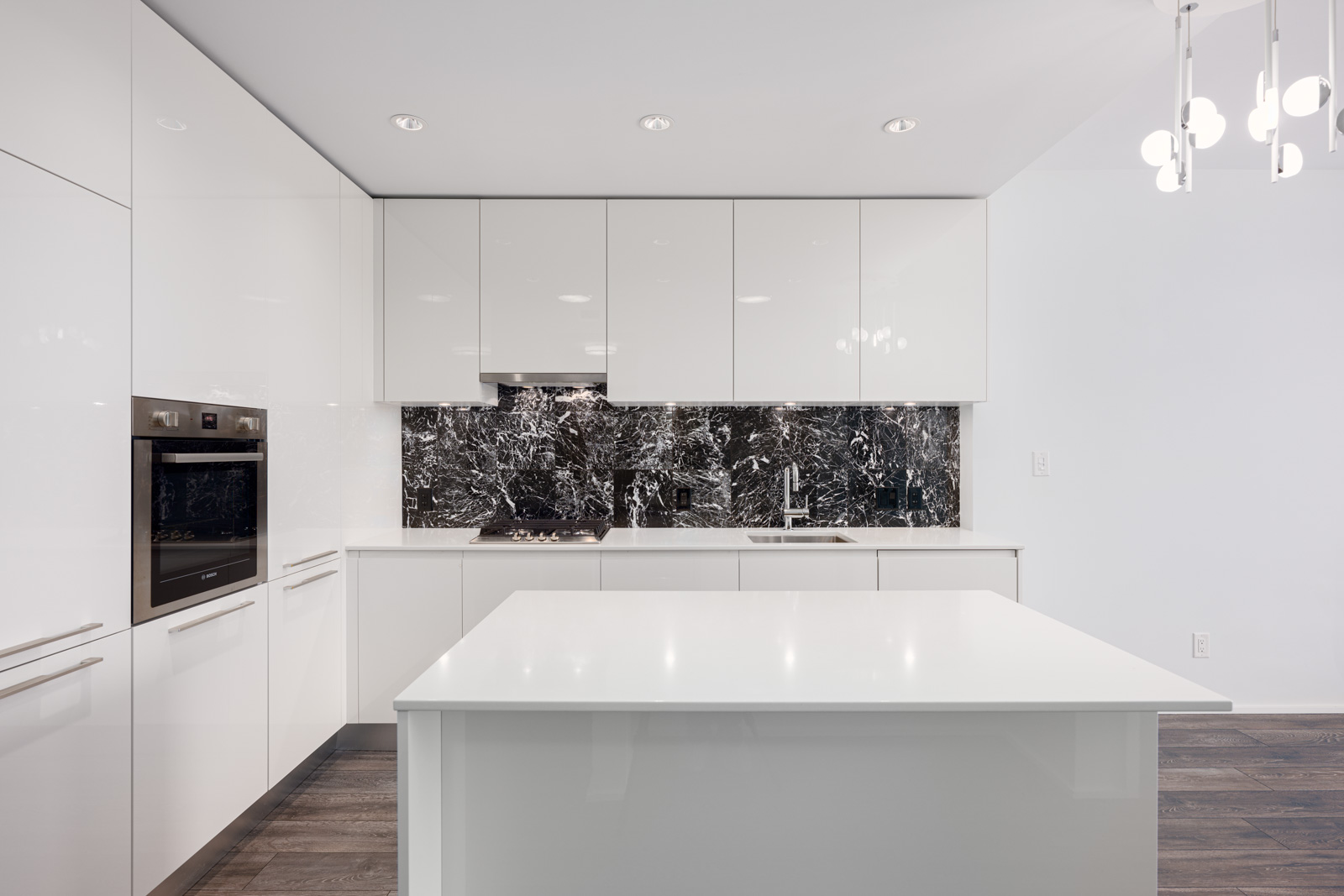 Modern kitchen with glossy white cabinets, marble backsplash, built-in oven, stovetop, sink, and a white island under recessed and pendant lighting.
