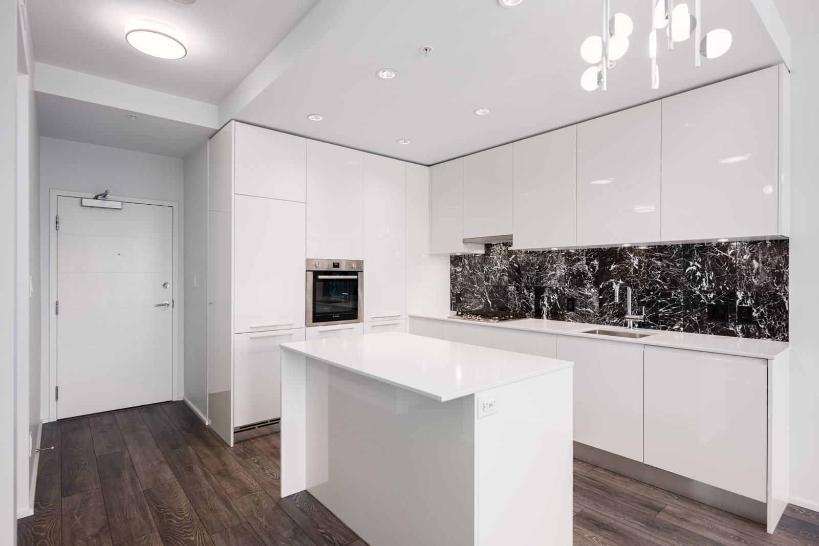 Modern kitchen with white cabinets, black marble backsplash, built-in oven, island countertop, and dark wood flooring under bright ceiling lights.