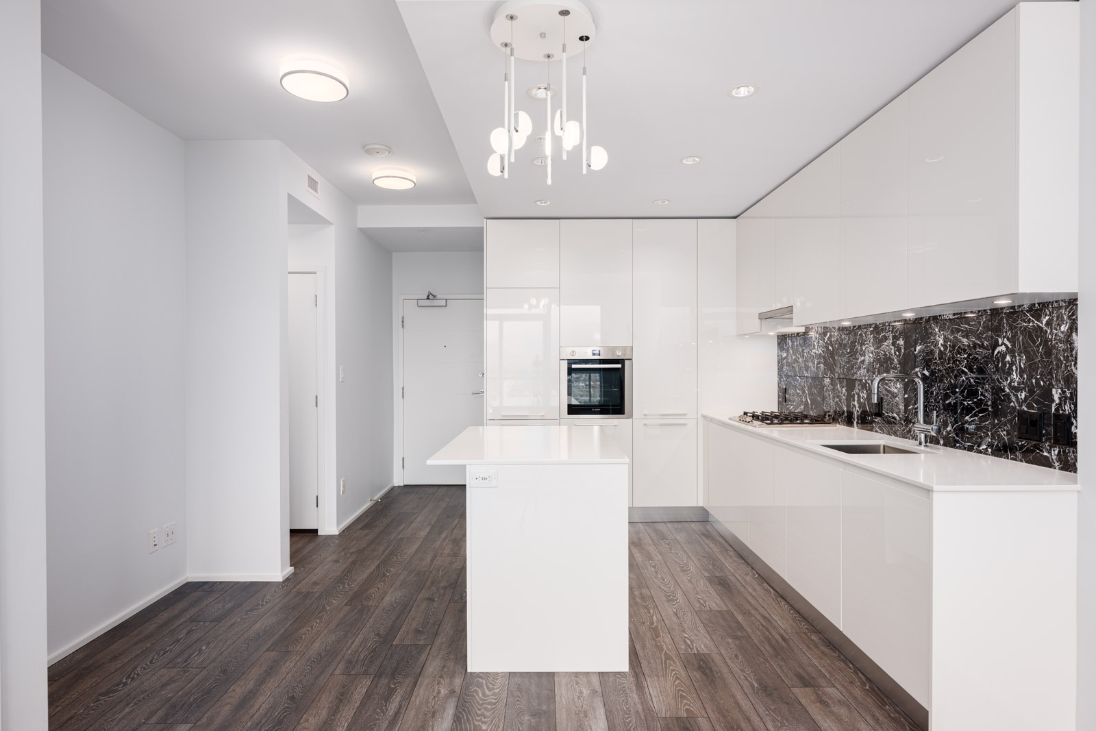 Modern kitchen with glossy white cabinets, dark marble backsplash, built-in oven, island, stainless steel sink, and wood flooring under bright ceiling lights.