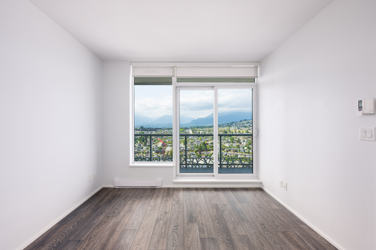 Empty room with light gray walls, dark wood flooring, and a large windowed door opening to a balcony with a mountain and city view.