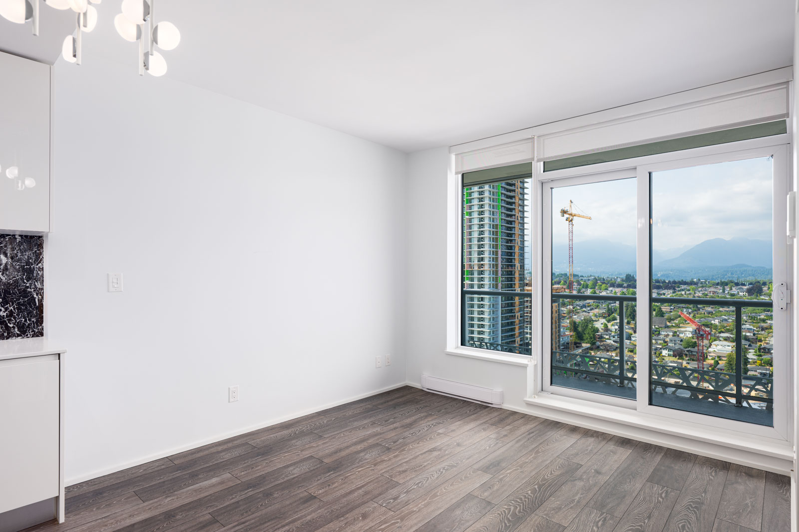 Unfurnished modern apartment room with large window and sliding glass door leading to a balcony overlooking a cityscape with high-rises, a crane, and mountains in the distance.