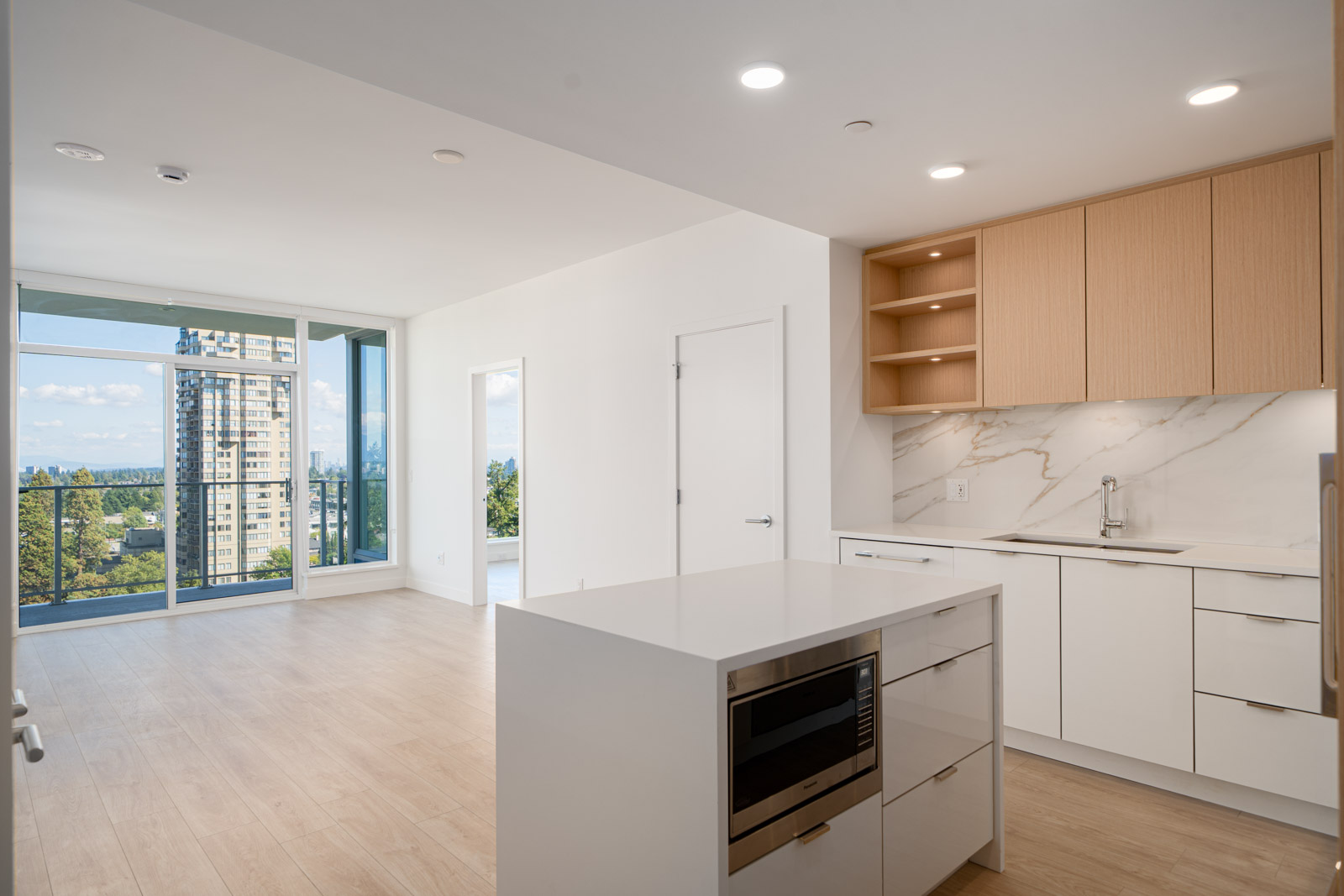 Modern apartment interior with a white kitchen island, built-in microwave, light wood cabinets, and large windows offering a city and forest view.