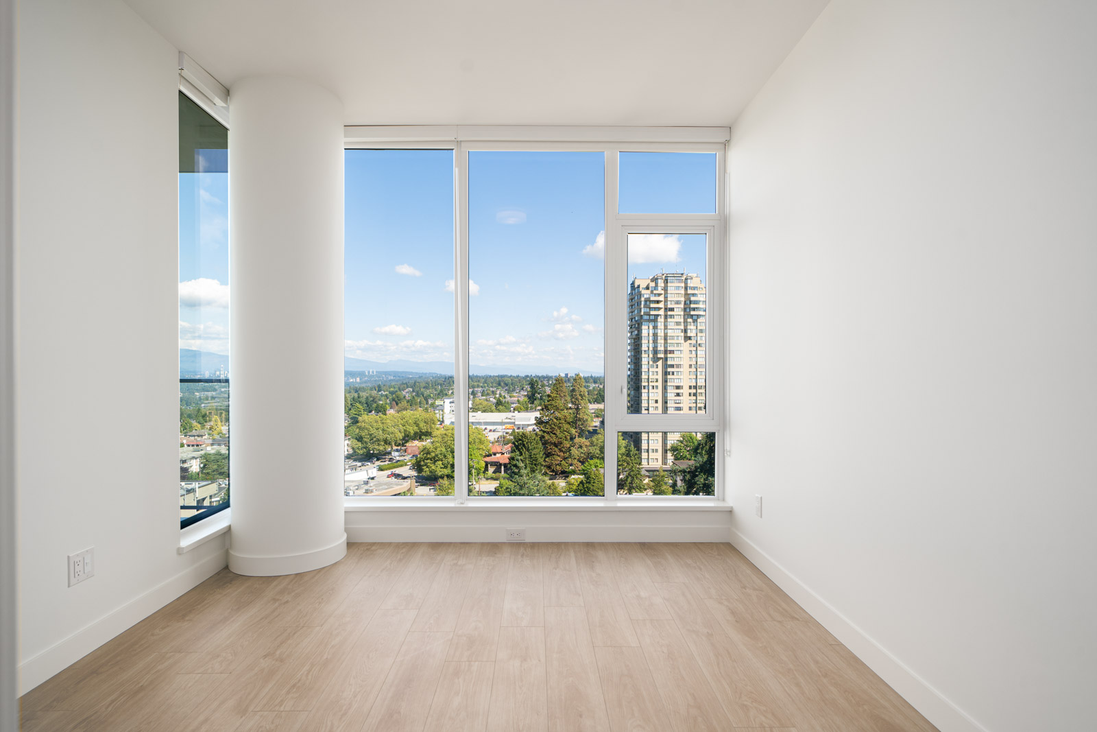 Empty room with light wood flooring, white walls, and large floor-to-ceiling windows offering a city and nature view.