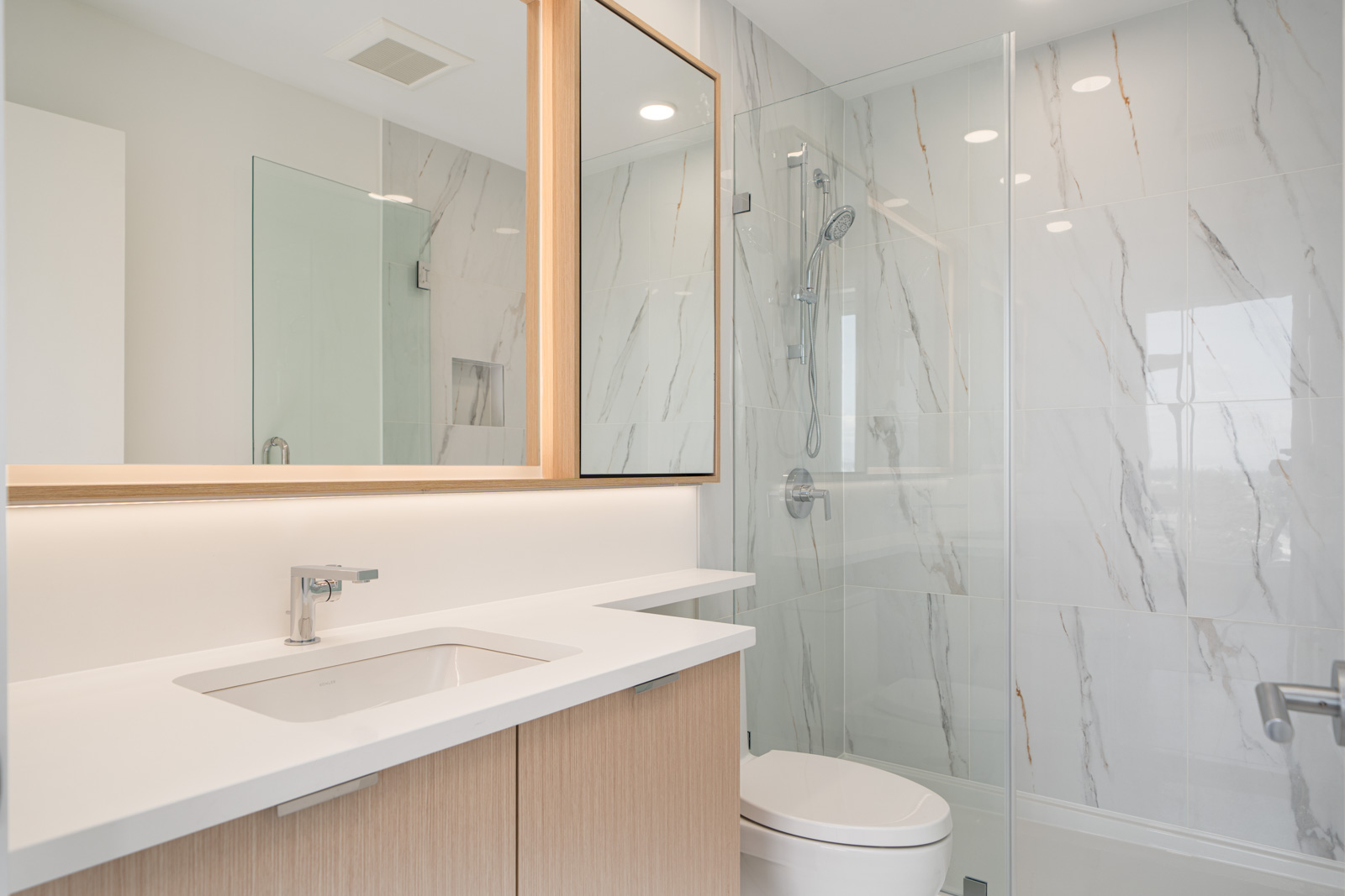 Modern bathroom with a white sink, light wood vanity, large mirror, glass shower enclosure, and white marble walls with gray veining.