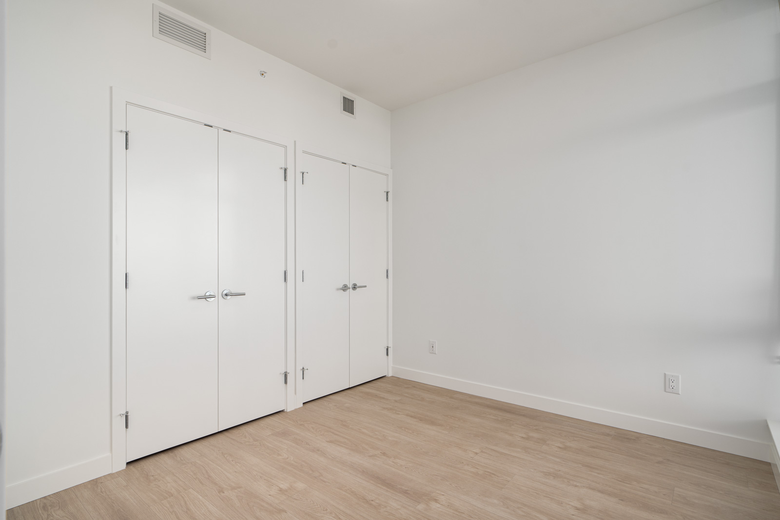 Empty room with light wood flooring, white walls, two closed closet doors, and a closed single door.