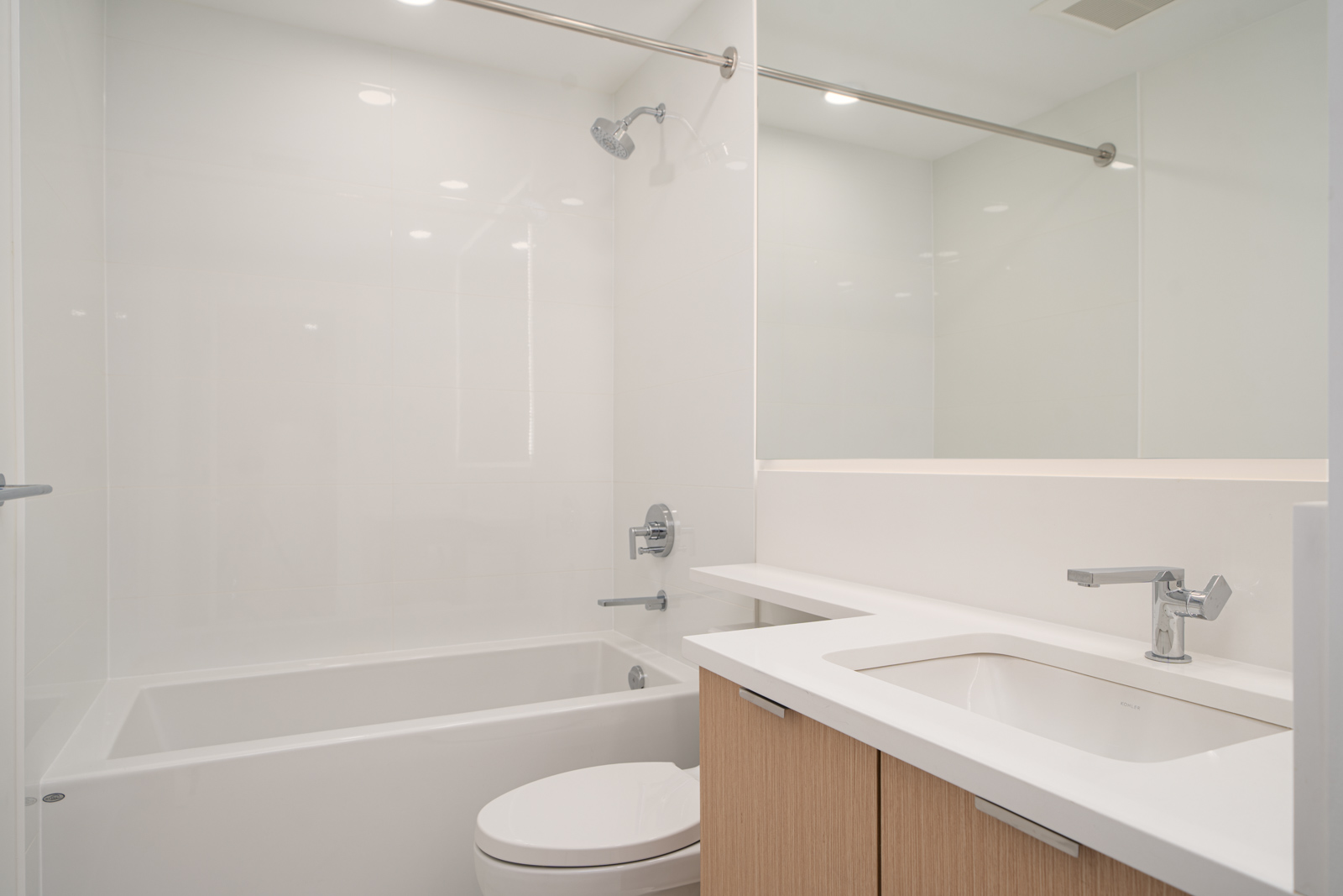 A modern bathroom with a white bathtub and shower, a toilet, a sink with a chrome faucet, and light wood vanity cabinets under a wide countertop.