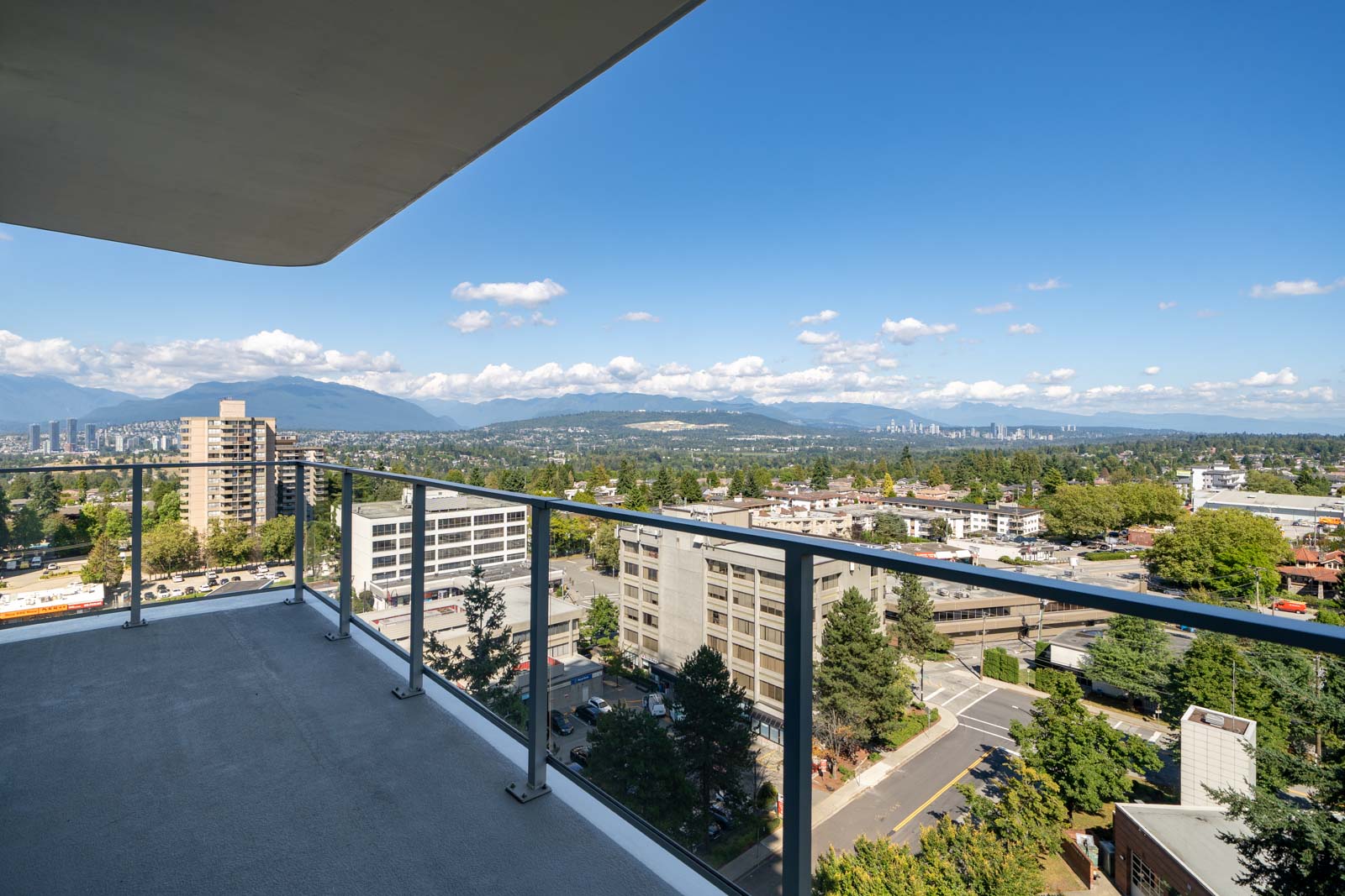 View from a high-rise balcony overlooking city buildings, green areas, and distant mountains under a clear blue sky.