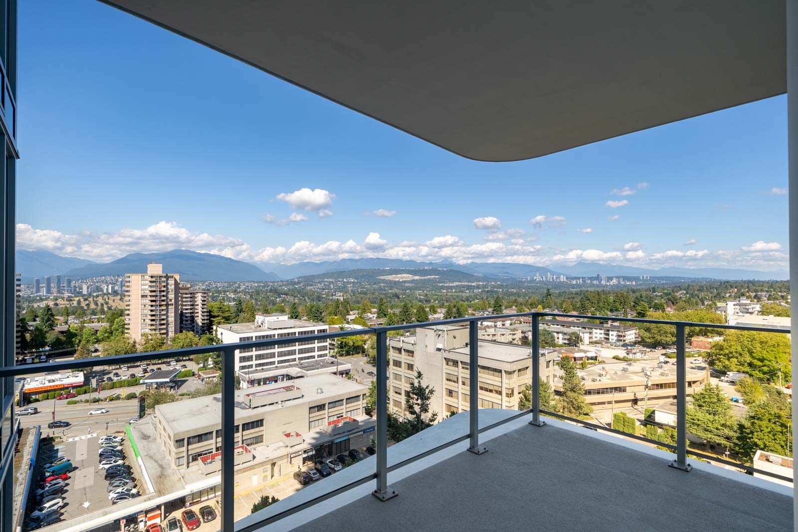View from a high balcony showing city buildings, trees, distant mountains, and a clear sky with scattered clouds.