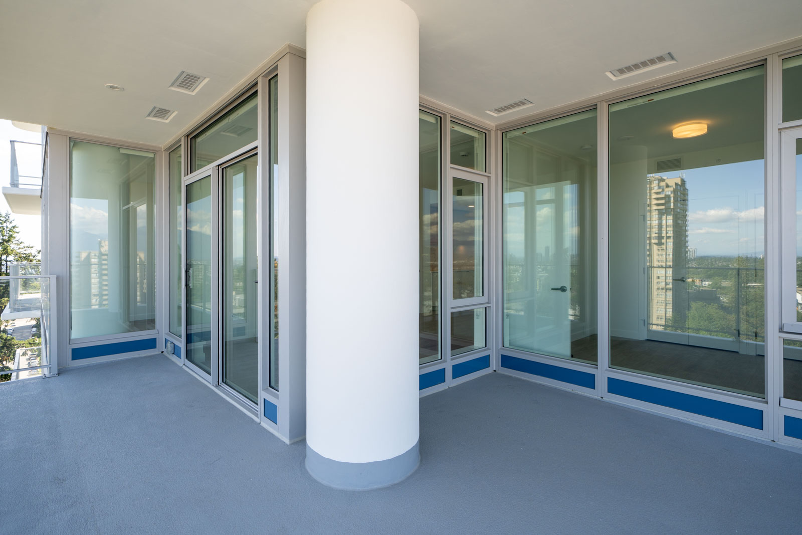 Modern apartment balcony with a large white pillar, glass doors, and windows overlooking city buildings and greenery in the background.