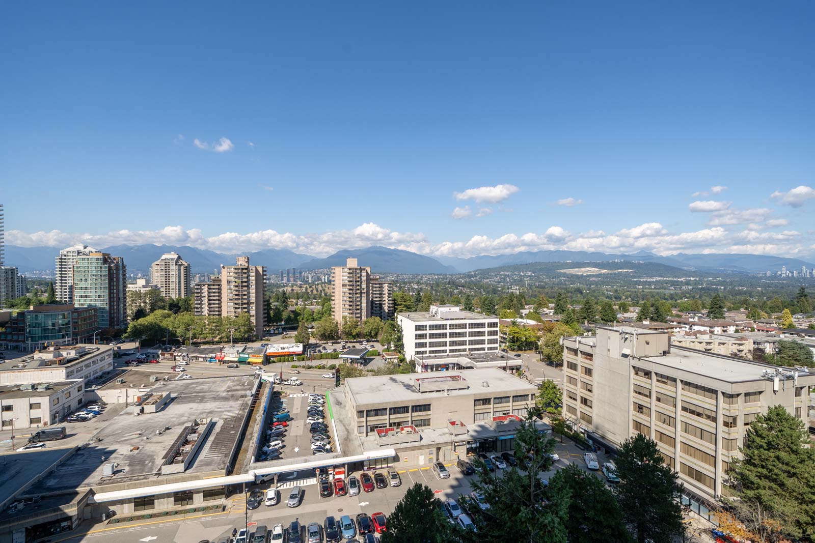 Aerial view of a cityscape with mid-rise buildings, parking lots, and distant mountains under a clear blue sky with scattered clouds.