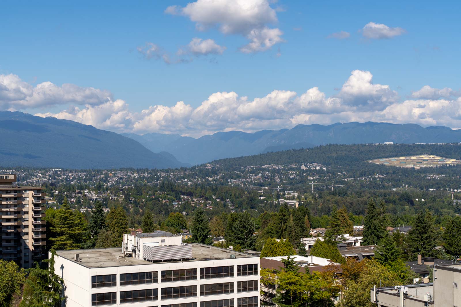 A cityscape with mid-rise buildings in the foreground, dense greenery, and distant mountains under a partly cloudy sky.