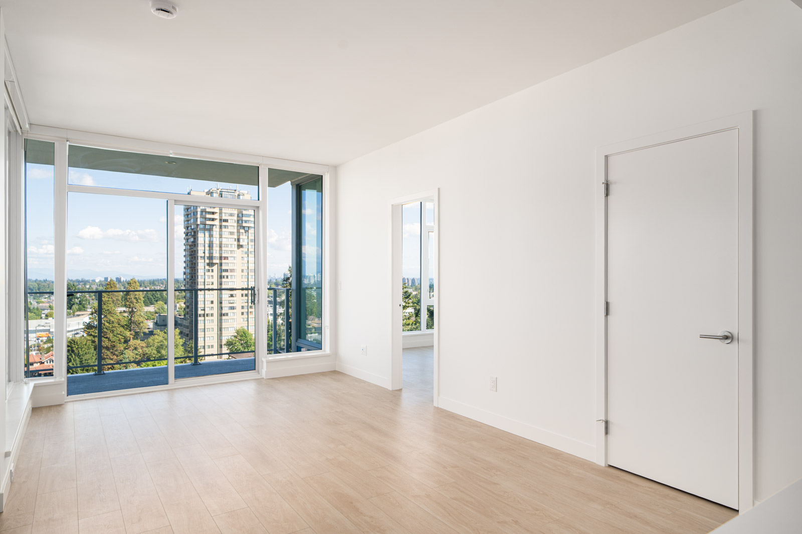 Unfurnished modern apartment room with light wood flooring, white walls, large windows, glass balcony door, and city view.