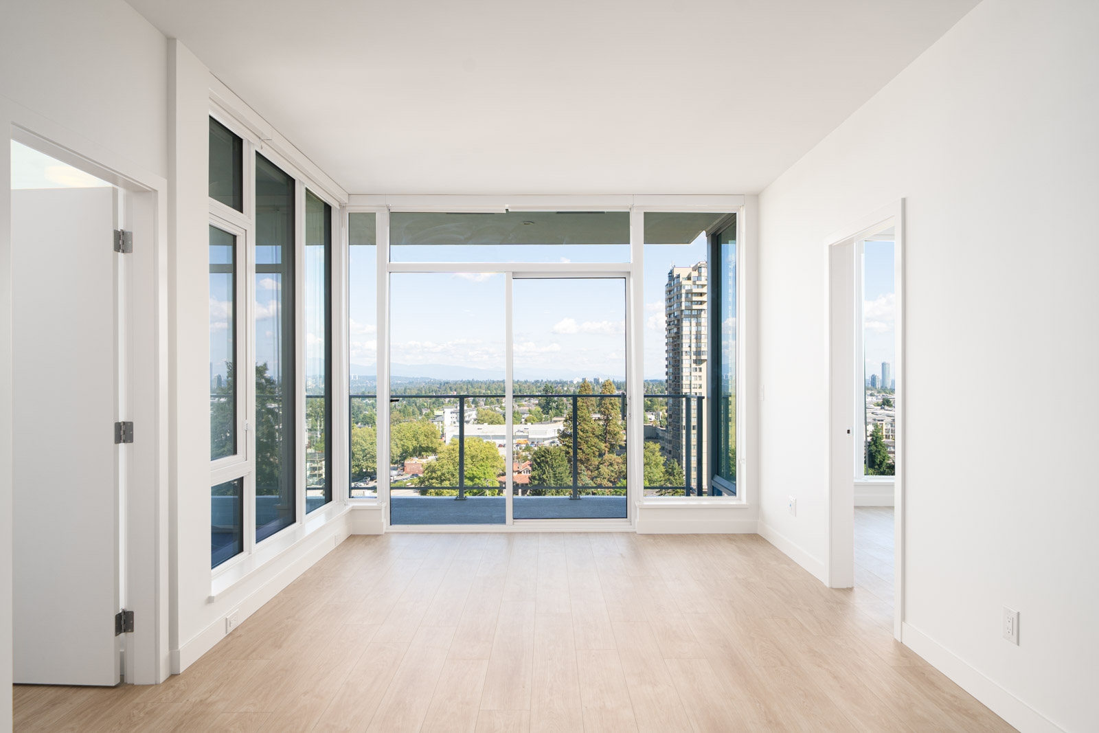 Bright, empty modern apartment room with light wood flooring, large floor-to-ceiling windows, sliding glass door to balcony, and cityscape view in the background.