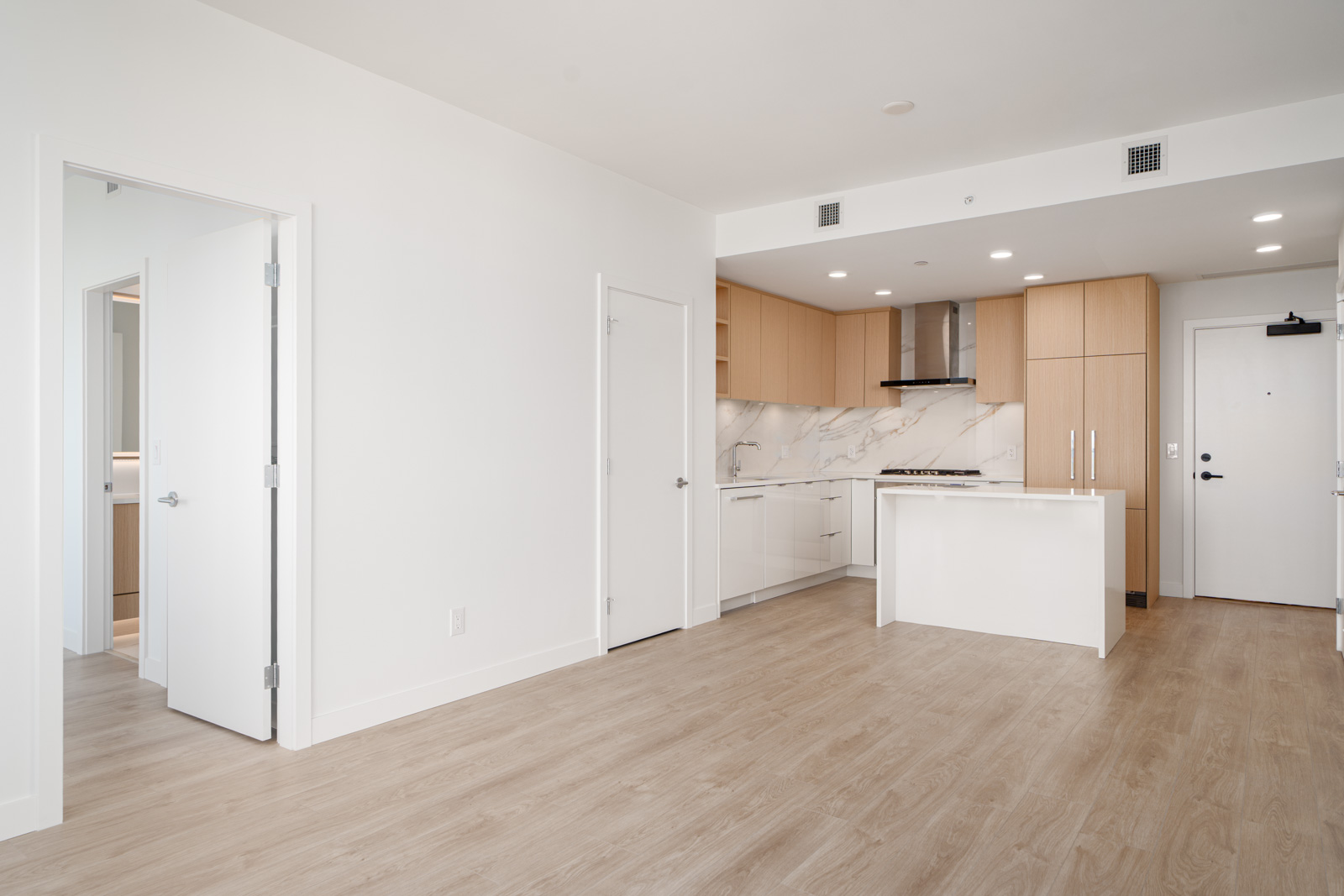 Modern, unfurnished apartment with wood floors, a white and wood kitchen, marble backsplash, two open doors, and recessed lighting.
