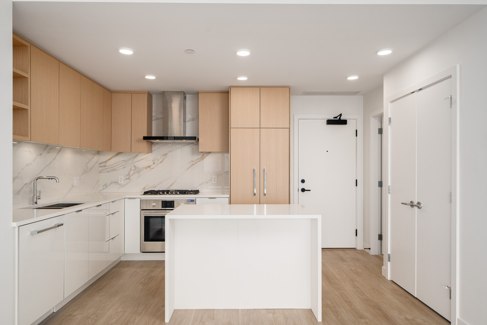 Modern kitchen with light wood cabinets, white countertops, marble backsplash, a central island, built-in oven, and stainless steel range hood. Wood flooring and white walls throughout.