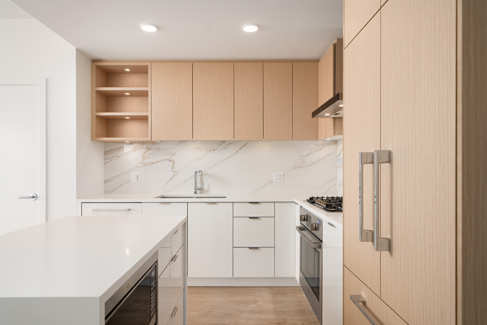 Modern kitchen with light wood cabinets, white countertops, marble backsplash, built-in oven, gas stove, and a kitchen island with a microwave.