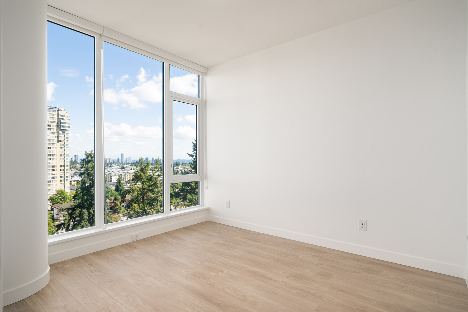 Empty corner of a modern room with light wood flooring, white walls, and large floor-to-ceiling windows overlooking a city skyline and trees.