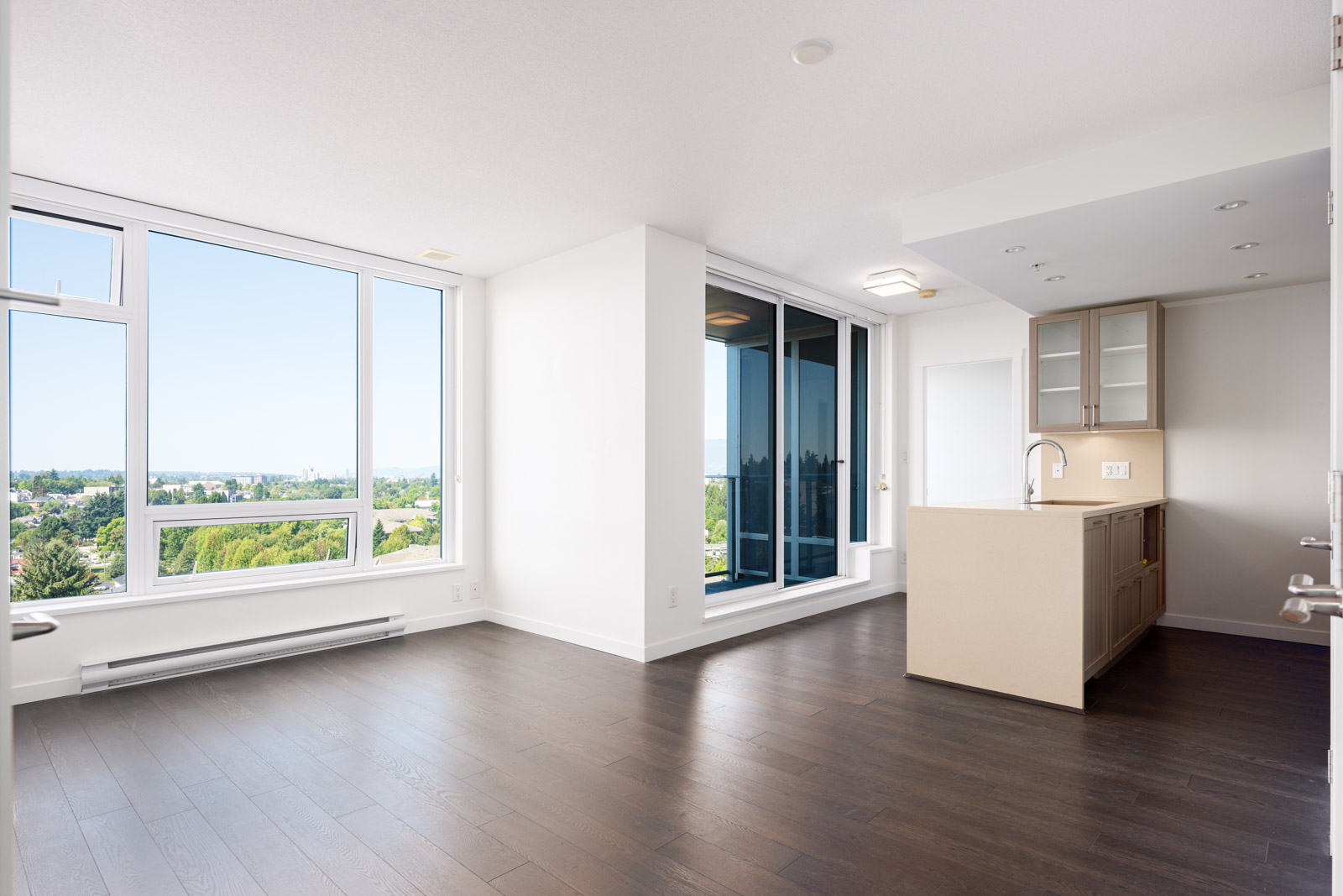 Bright, unfurnished apartment with large windows, hardwood floors, an open living area, and a modern kitchen island. Balcony seen through glass doors, offers a view of greenery outside.