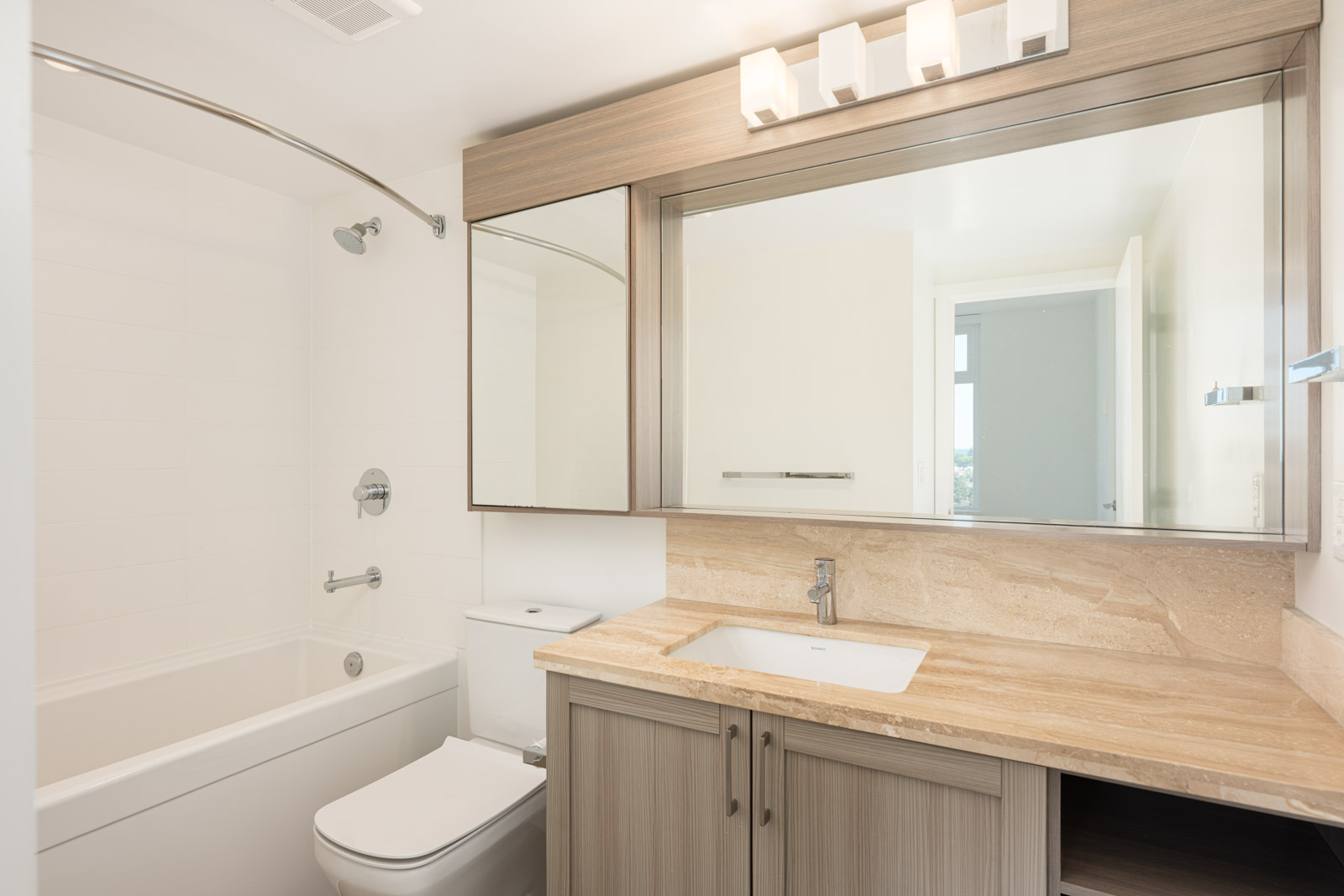 Modern bathroom with a bathtub-shower combo, toilet, large mirror, wood cabinetry, and beige marble countertop under bright ceiling lights.