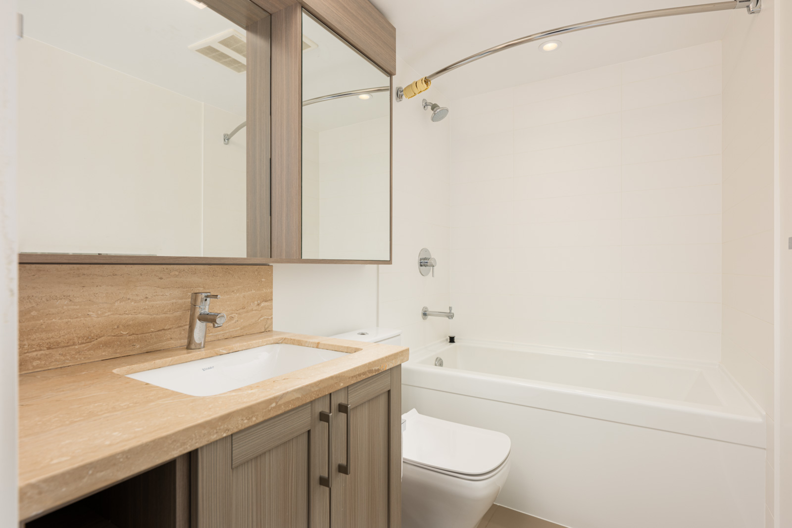 Modern bathroom with a wood vanity, beige countertop, rectangular sink, toilet, and a white bathtub with a curved shower rod.