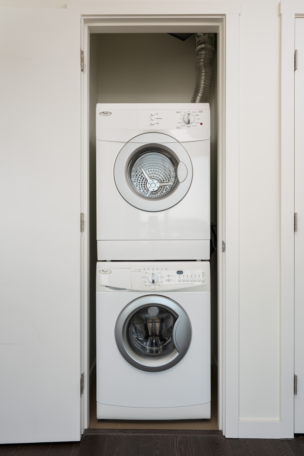 A stacked washer and dryer unit is installed in a small, open closet with white walls and dark flooring.