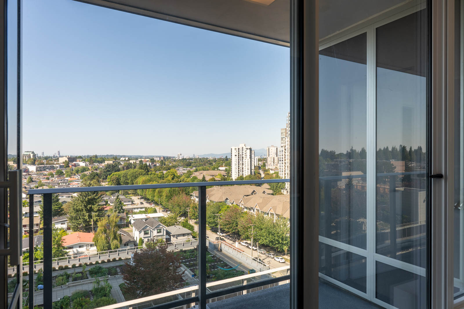 A cityscape view from a balcony showing residential buildings, trees, and a clear blue sky, seen through a glass door.