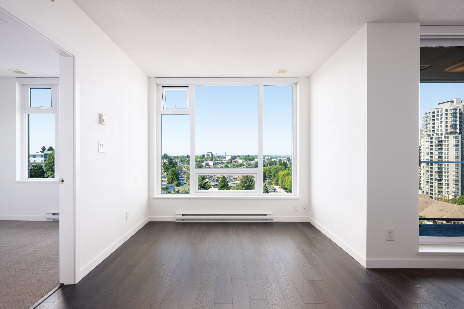 Empty bright room with large windows, dark wood flooring, and cityscape view; adjacent room partially visible on the left.