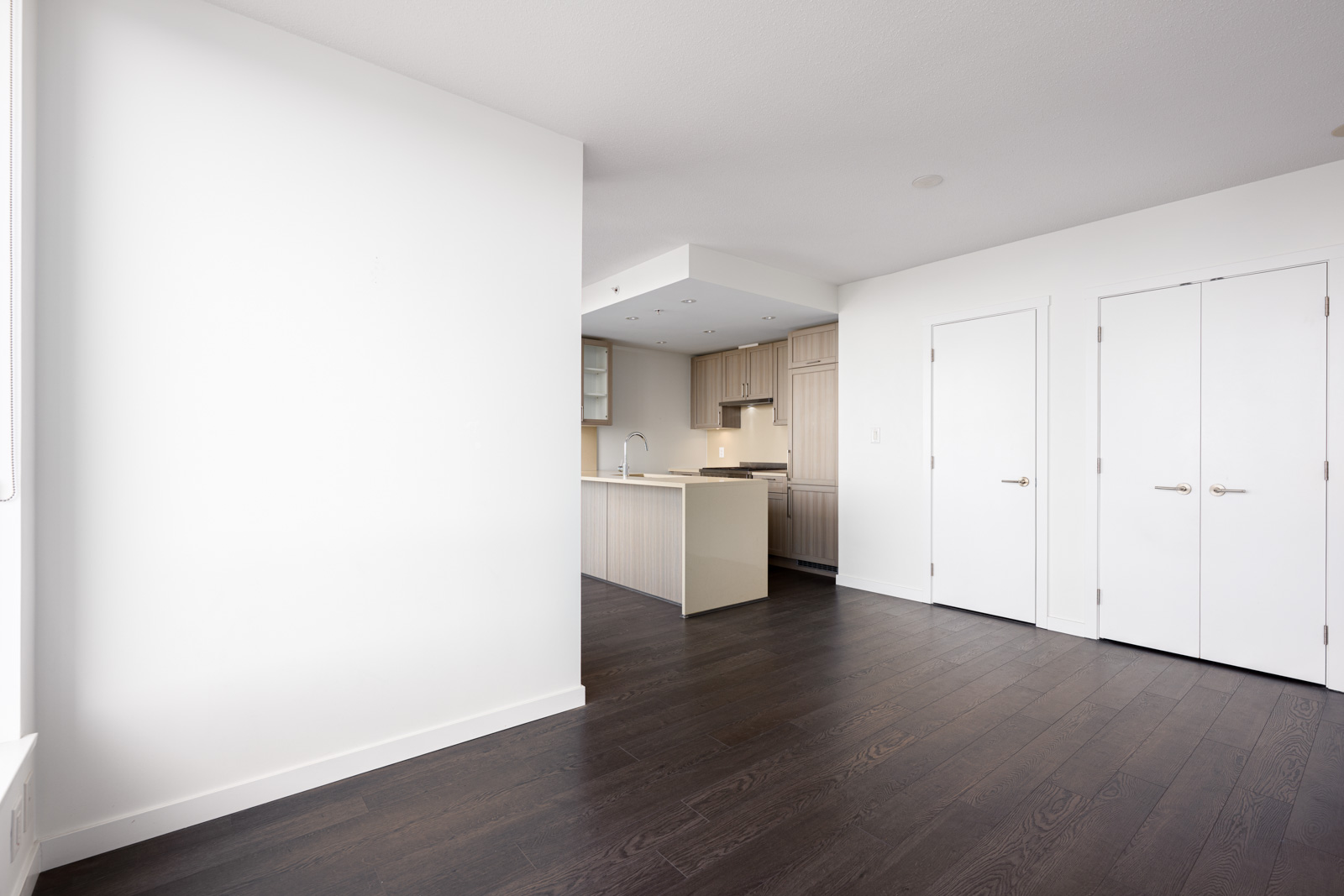 Empty modern apartment interior with dark wood floors, white walls, a small kitchen area with light cabinets, and two closed white doors.