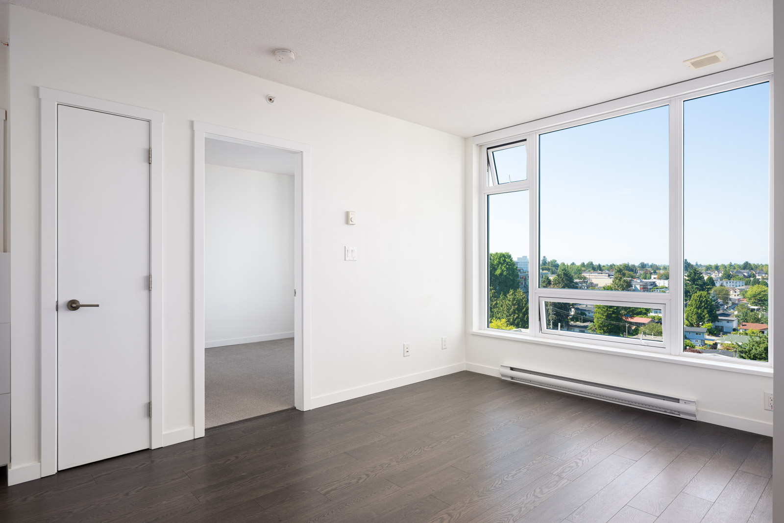 Bright empty room with white walls, dark wood flooring, a white door, an open doorway, and large windows overlooking a green outdoor landscape under a clear sky.