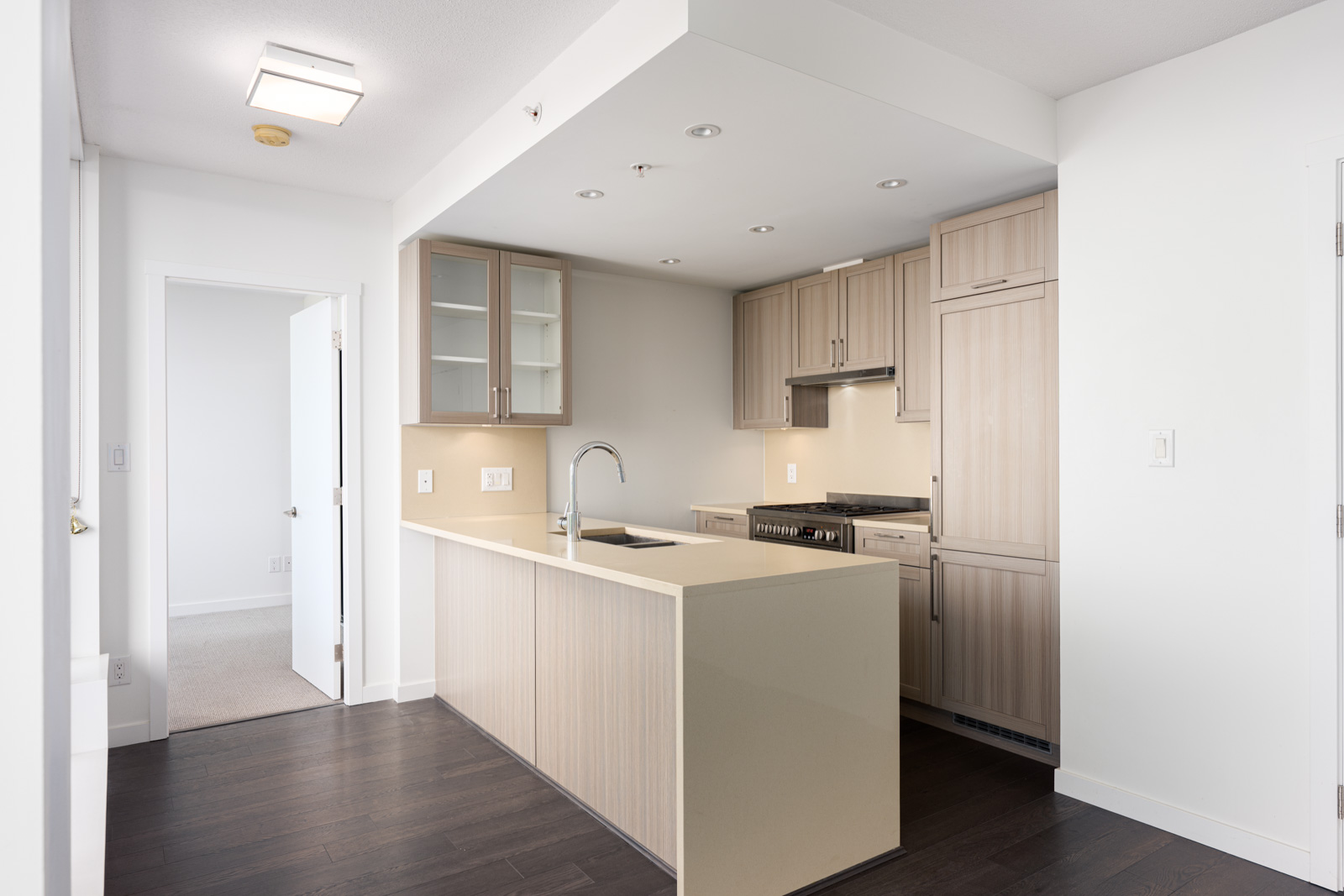 Modern kitchen with light wood cabinets, built-in appliances, a center island with a sink, and dark hardwood flooring, adjacent to a carpeted room through an open doorway.