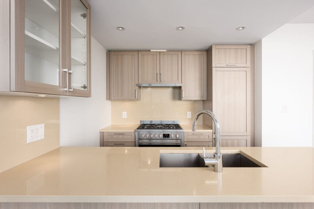 Modern kitchen with light wood cabinets, stainless steel stove, built-in refrigerator, beige countertops, and a sink with a gooseneck faucet on the island.
