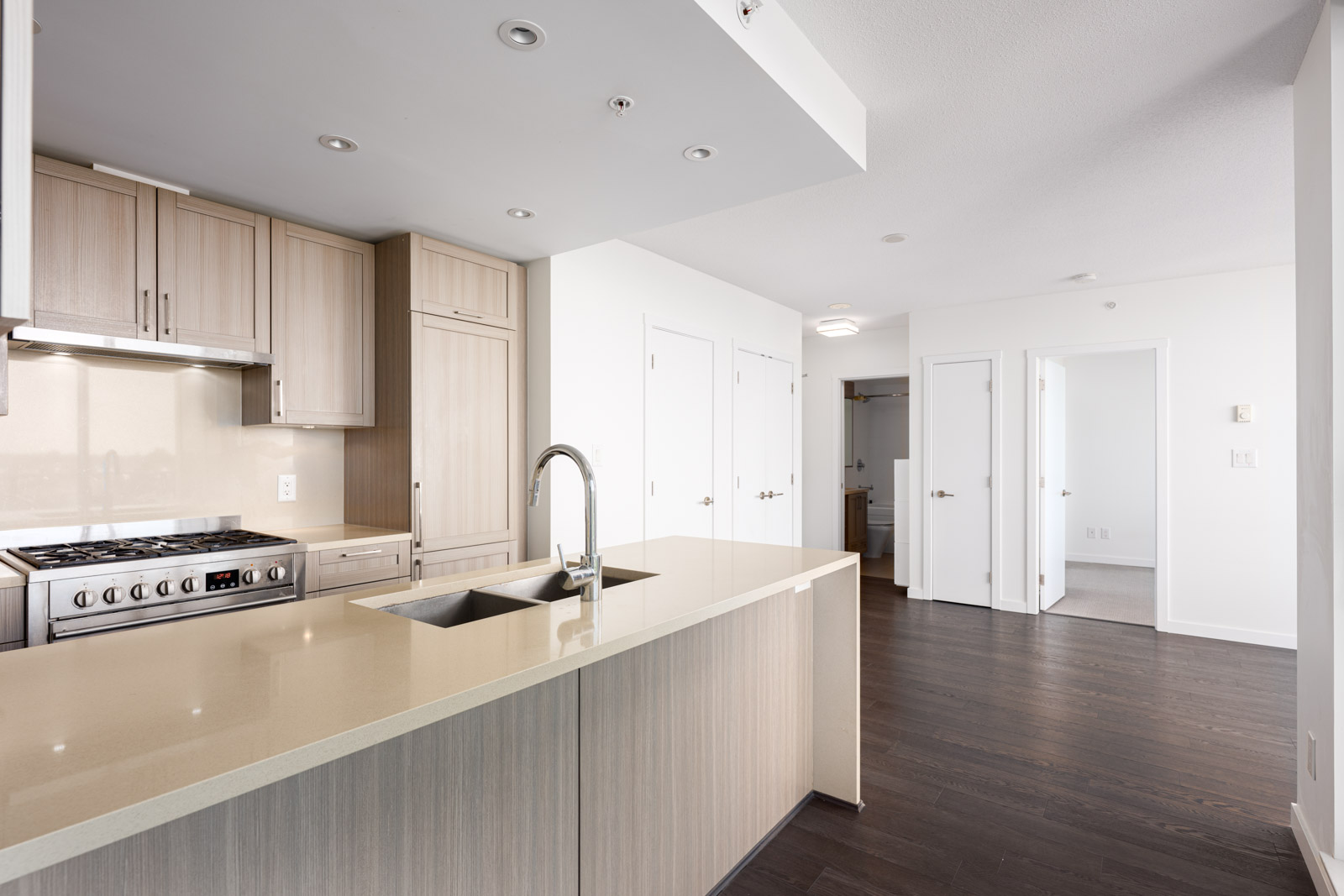 Modern kitchen with light wood cabinets, stainless steel appliances, a double sink island, and dark hardwood floors, opening into a bright hallway and adjoining rooms.
