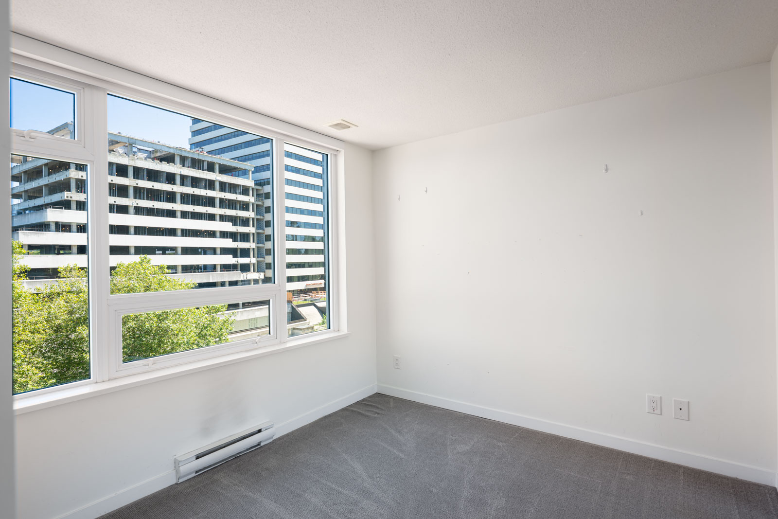 Empty room with white walls, grey carpet, and a large window showing a view of modern office buildings and trees outside.