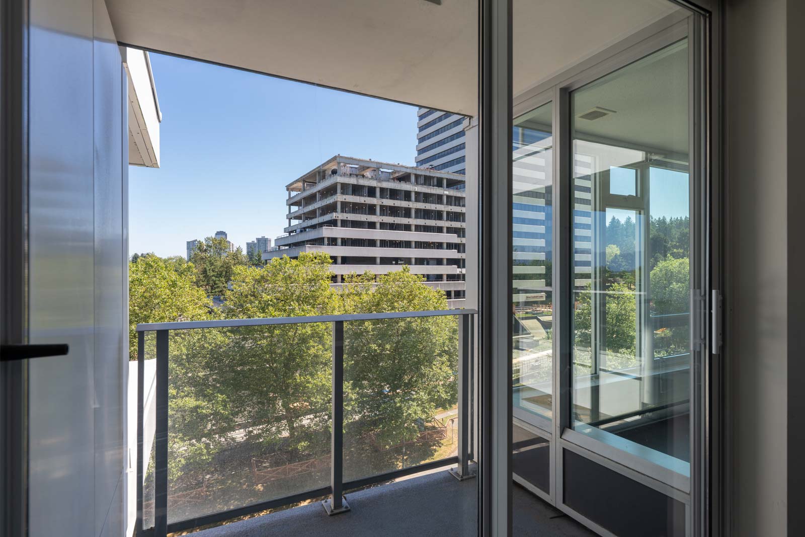 View from a glass balcony showing trees, an under-construction building, and cityscape in the background on a sunny day.