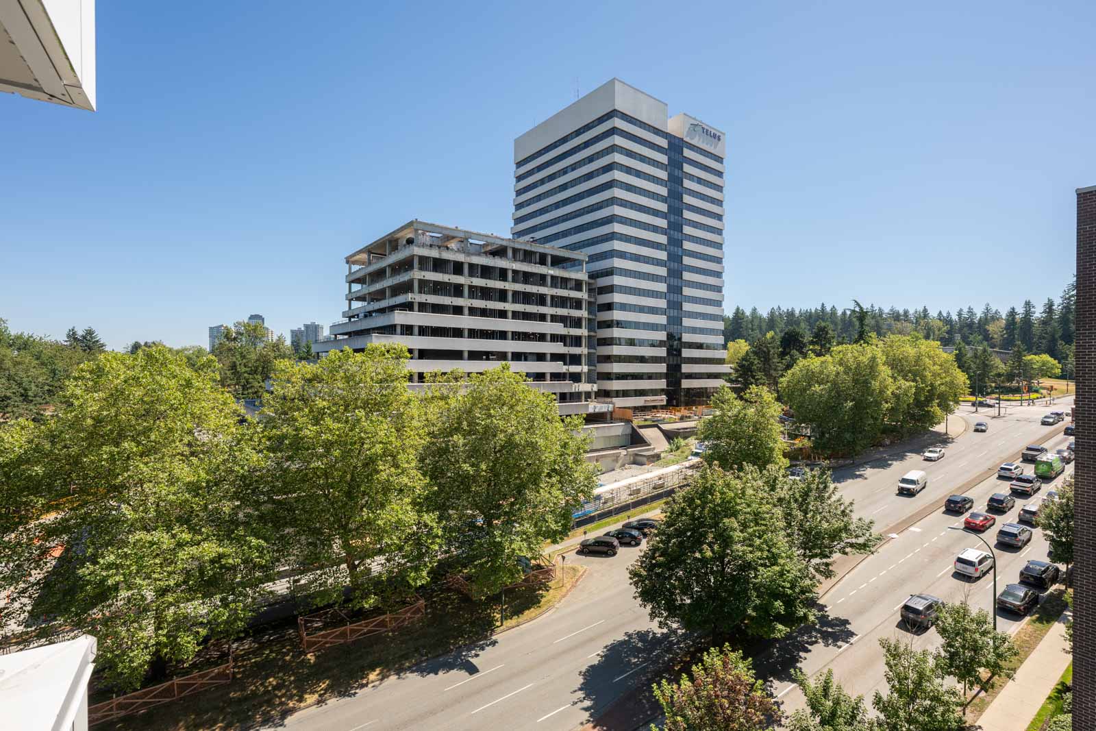 A multi-story building under demolition stands beside a tall office tower, surrounded by trees and a road with light traffic on a clear, sunny day.
