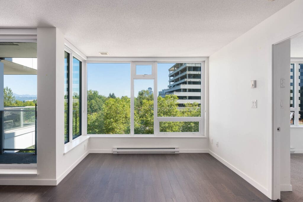 Unfurnished apartment room with large windows, dark wood flooring, and a view of trees and modern buildings outside.