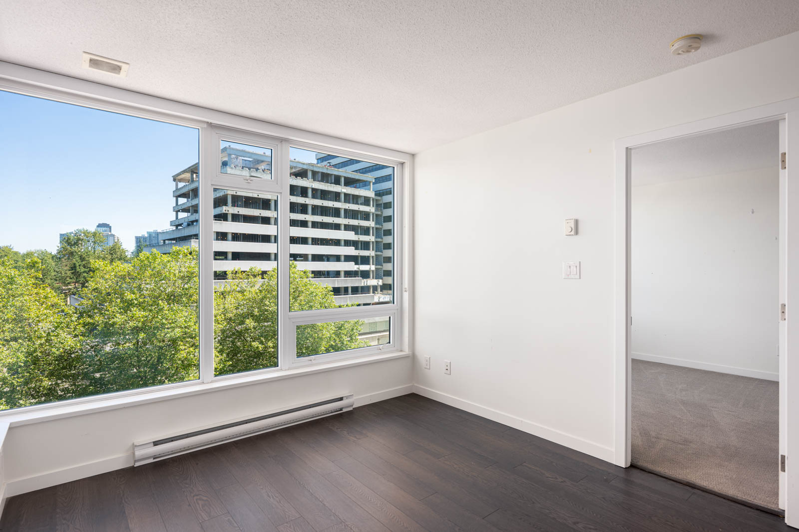 Bright room with large windows, dark wood flooring, white walls, and a view of trees and a modern building outside; doorway leads to a carpeted adjacent room.