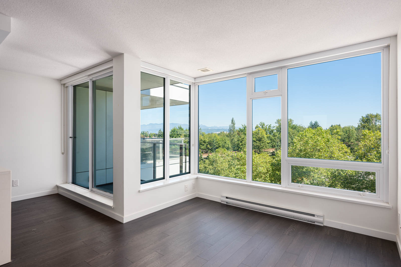 Bright, empty room with large floor-to-ceiling windows, dark wood flooring, and a view of trees and mountains outside.