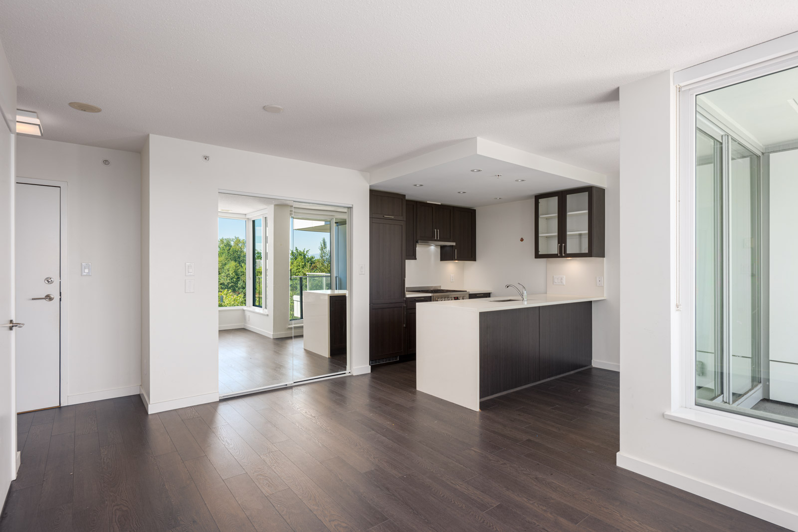 Modern, unfurnished apartment with dark wood floors, white walls, open kitchen with dark cabinets, large windows, and a glass-enclosed balcony with views of greenery.