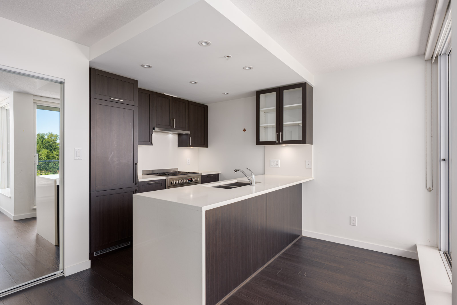 Modern kitchen with dark wood cabinets, white countertops, a built-in oven, and a sink island, adjacent to a window and sliding glass door.