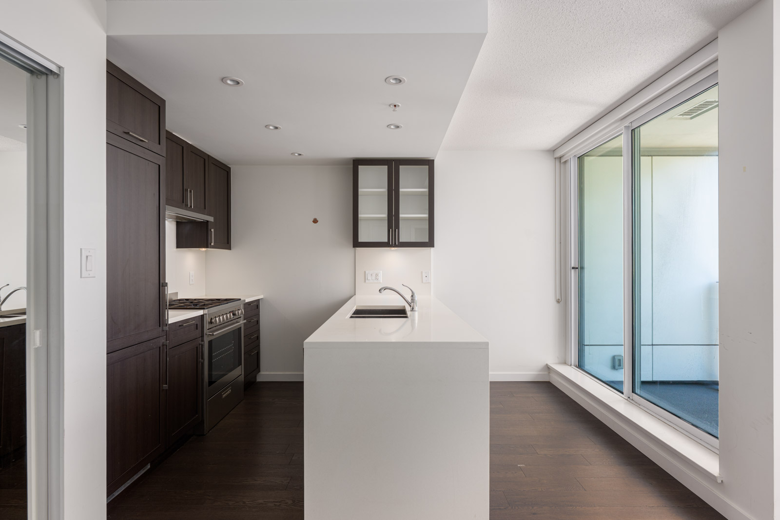 Modern kitchen with dark wood cabinets, white countertops, island sink, built-in stove, and large sliding glass doors leading to a balcony.