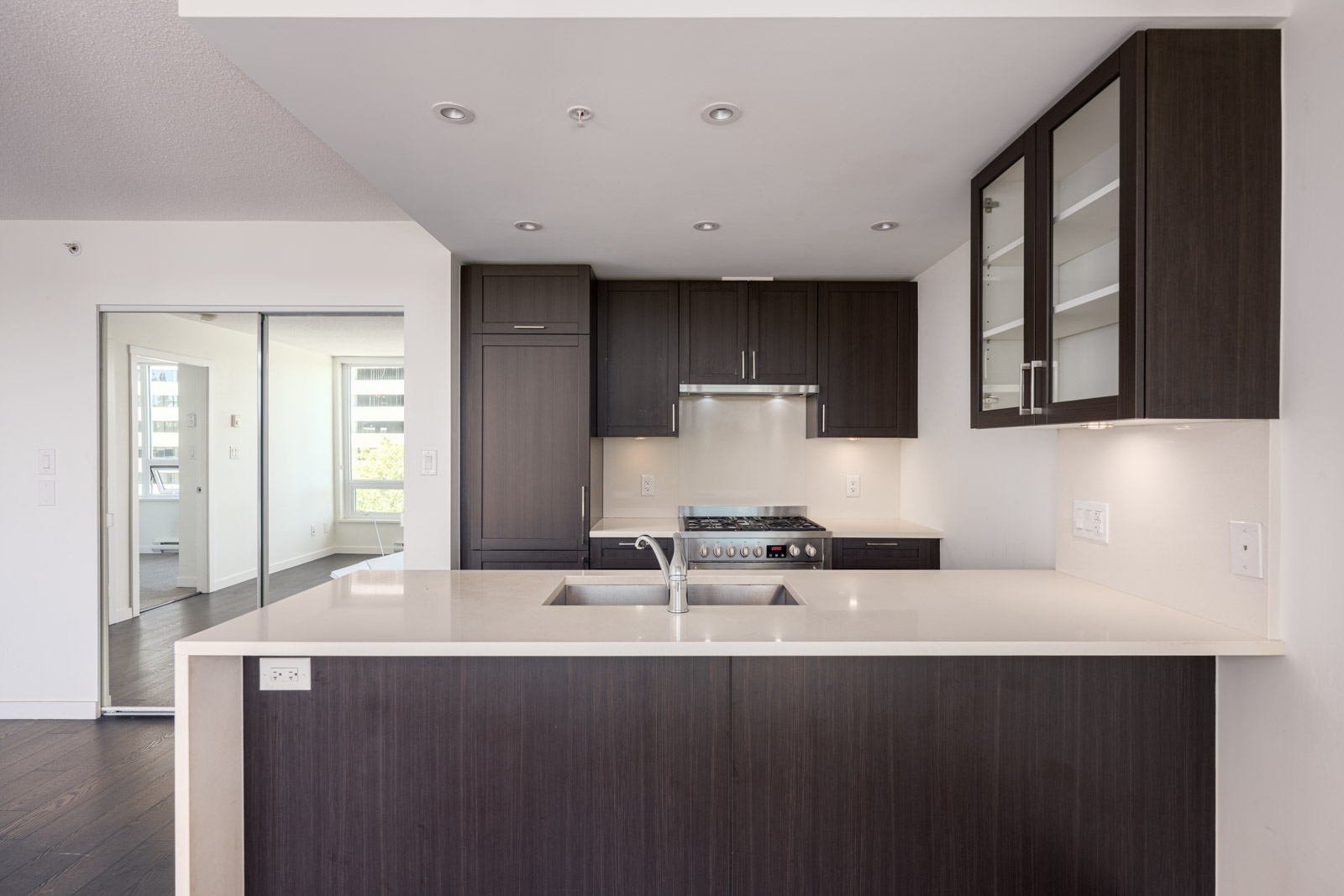Modern kitchen with dark wood cabinets, white countertops, built-in stove, sink, and glass-fronted upper cabinets; sliding glass door leads to another room.