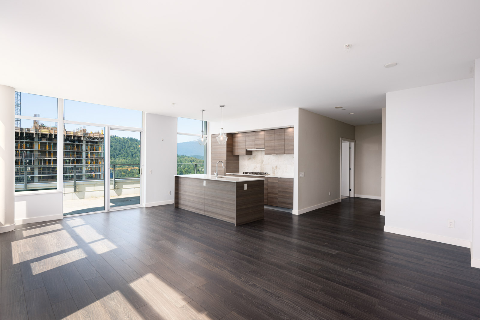 Modern empty apartment with dark wood floors, an open kitchen with island, and floor-to-ceiling windows overlooking a balcony and city view.