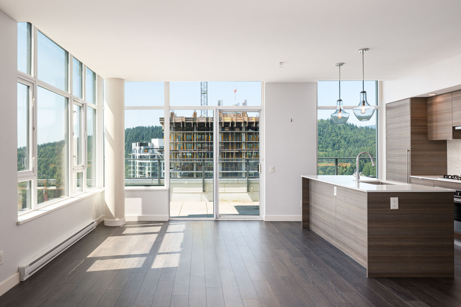 Modern empty apartment with large windows, dark wood flooring, open kitchen, and a view of a construction site and greenery outside.