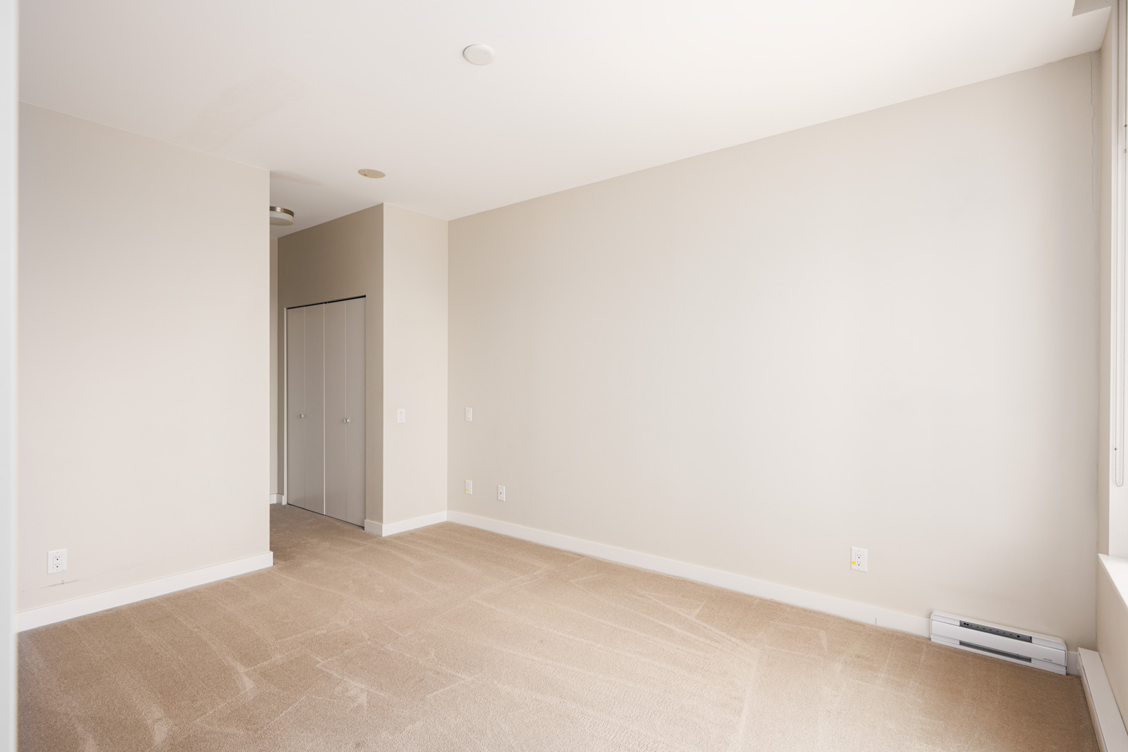 Empty beige carpeted room with white walls, a closet with double doors, and a window on the right letting in natural light.