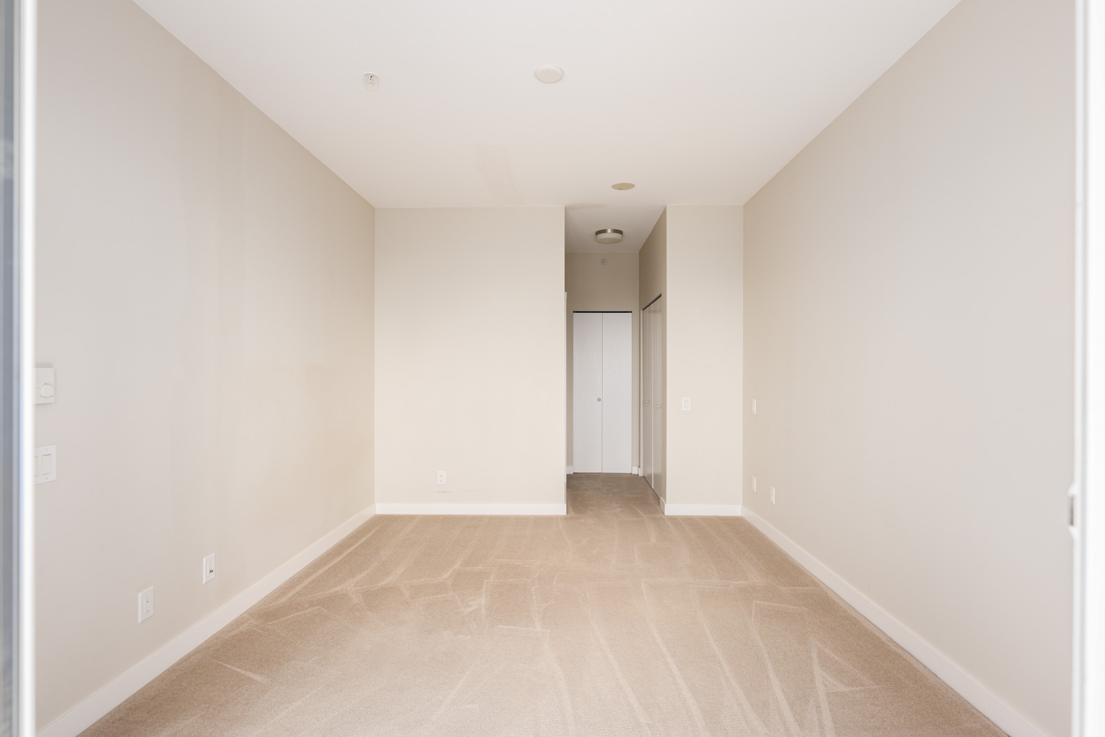 Empty room with beige carpet, white walls, and a small hallway leading to a closet with sliding white doors.