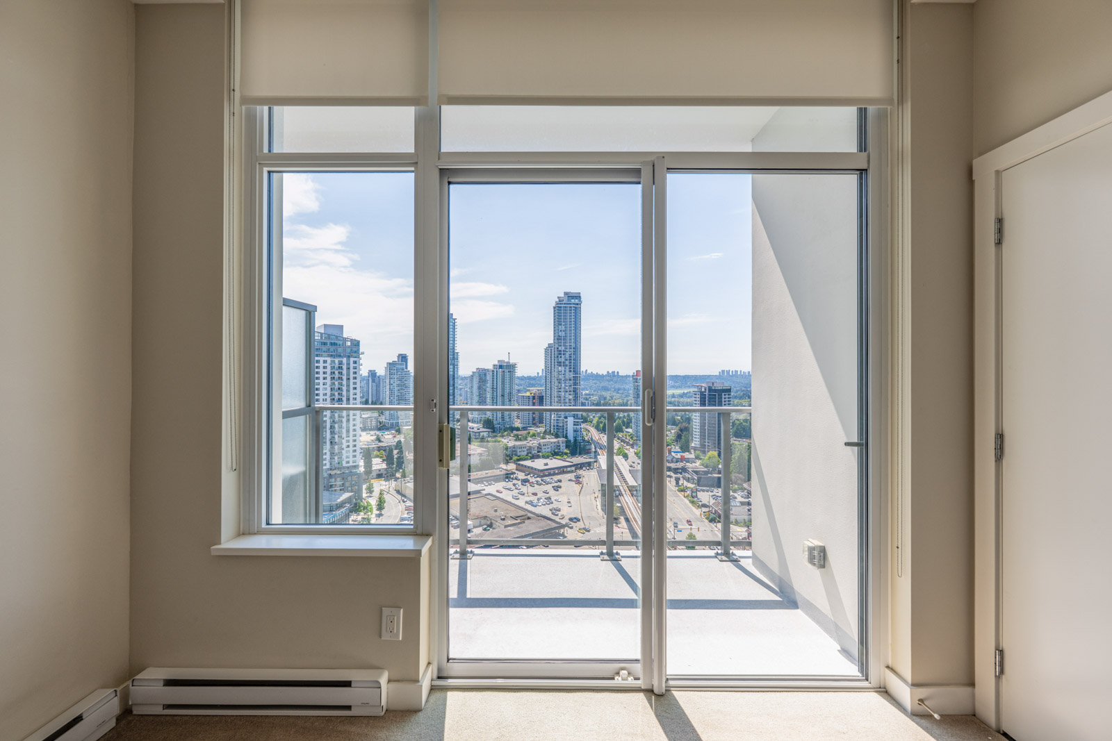 A high-rise apartment room with floor-to-ceiling windows and glass door opening to a balcony, overlooking a cityscape with tall buildings and clear sky.