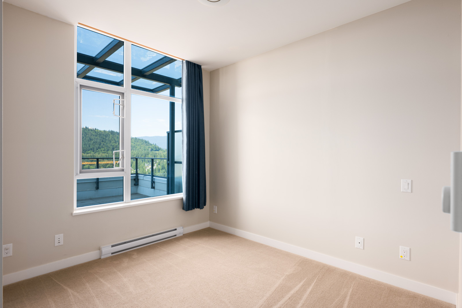 Empty room with beige carpet, white walls, a large window with a blue curtain, and a view of a balcony and greenery outside.