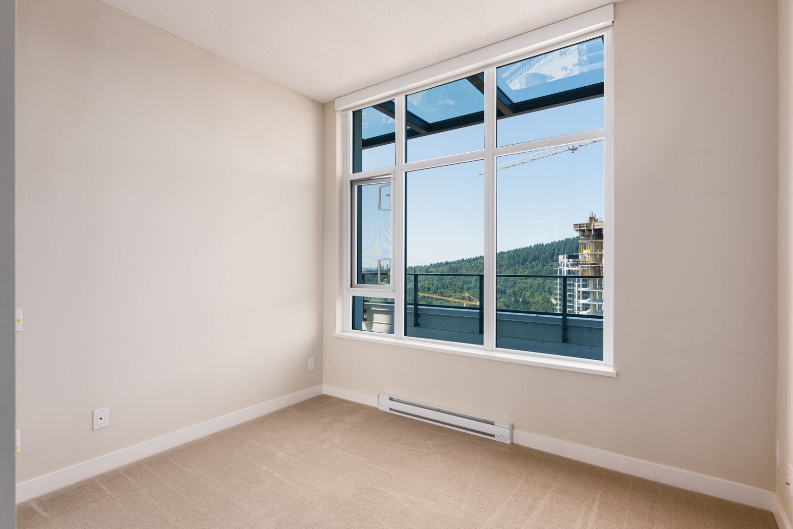 Empty room with beige carpet, light-colored walls, and large windows showing a view of a balcony, hills, and a construction crane outside.